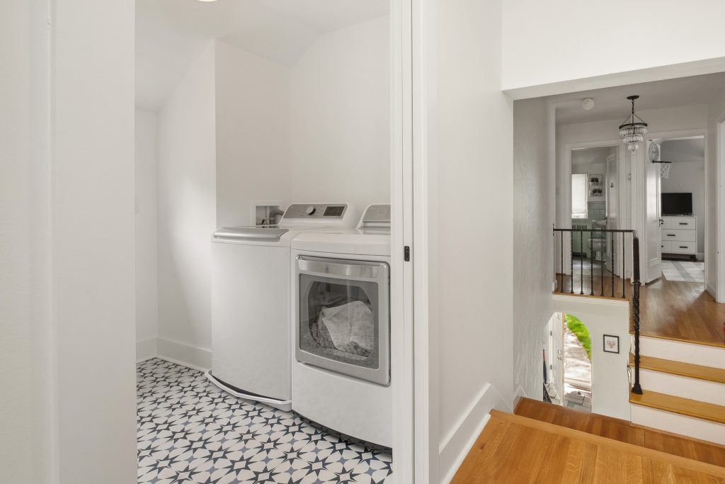 Compact laundry area with a washing machine and dryer next to a hallway with stairs and a railing, featuring patterned floor tiles and white walls.