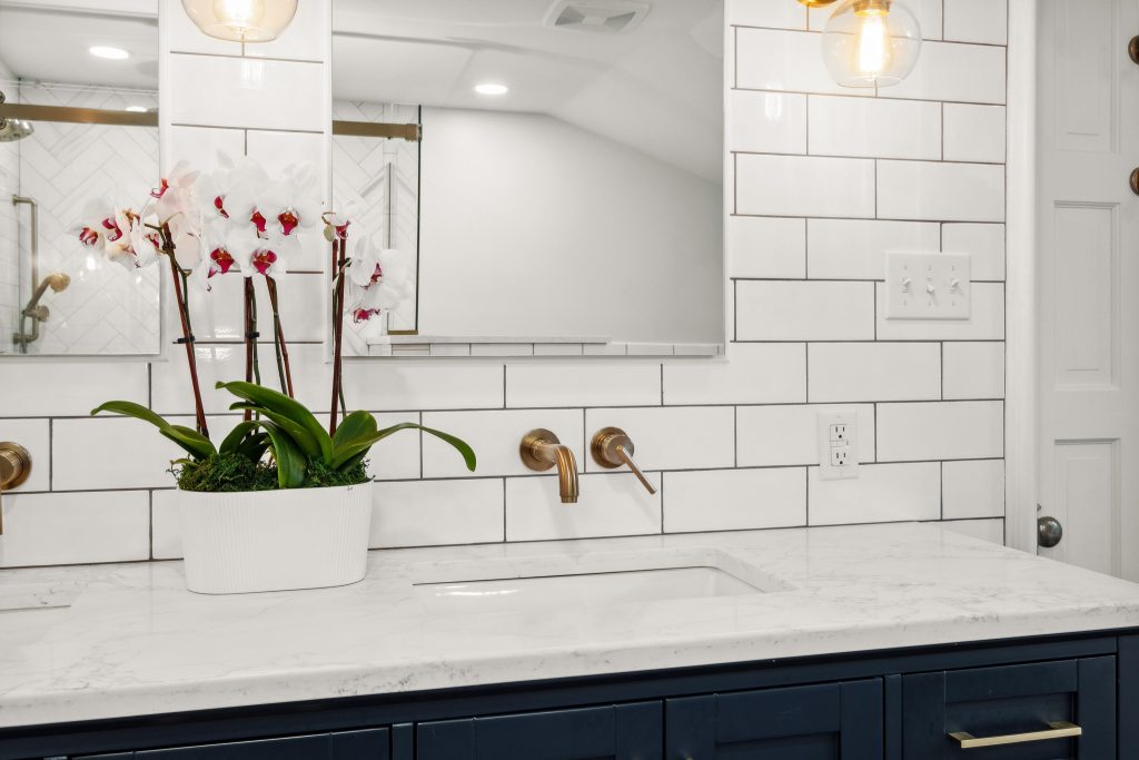 A modern bathroom features a white tiled wall, a white marble countertop, a rectangular mirror, and brass fixtures. A white pot with orchids is placed on the countertop.