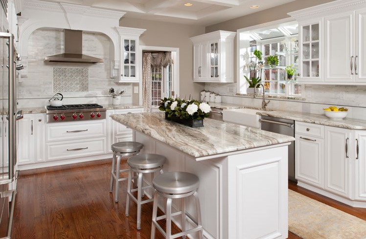 A modern kitchen with white cabinets, stainless steel appliances, a marble island with three metal stools, and hardwood floors, illuminated by natural light from large windows.