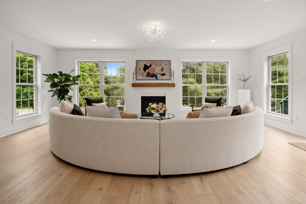 Spacious living room with a large curved sofa, centered fireplace, artwork above, and multiple windows letting in natural light. Some post-construction adjustments you'll want to budget for include window treatments.