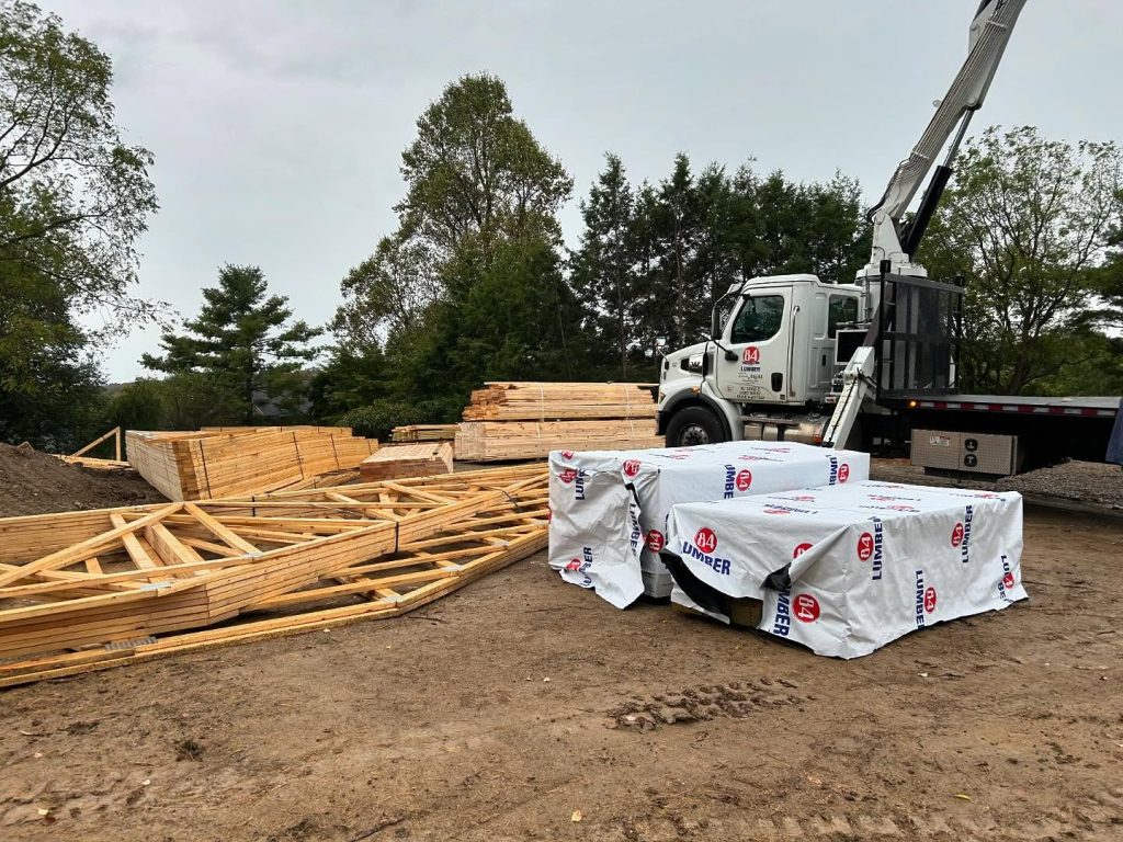 A truck unloads lumber at a construction site, with stacks of wooden beams and packaged materials on the ground. 