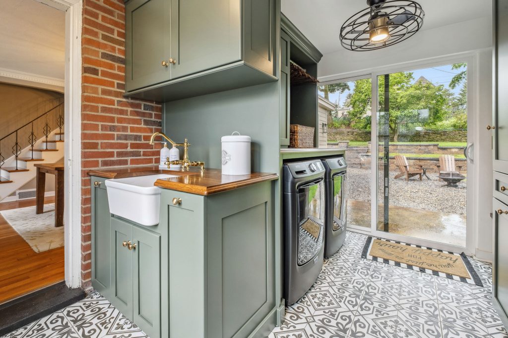 This laundry and mudroom addition has a white farmhouse utility sink, green cabinetry and washer and dryer units.