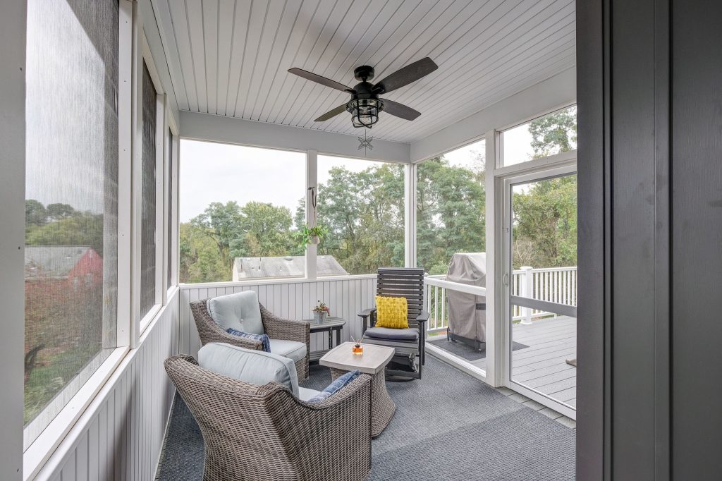 A screened-in porch with wicker chairs, a small table, and a ceiling fan offers a cozy spot to relax, reflecting thoughtful multi-generational home design with views of the deck and trees outside.