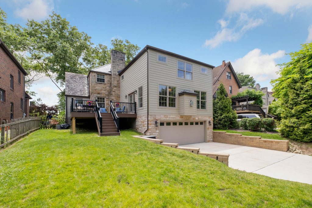 Two-story house built on a slope with stone and siding exterior, a raised deck, attached garage, driveway, and a grassy backyard bordered by fences and trees.