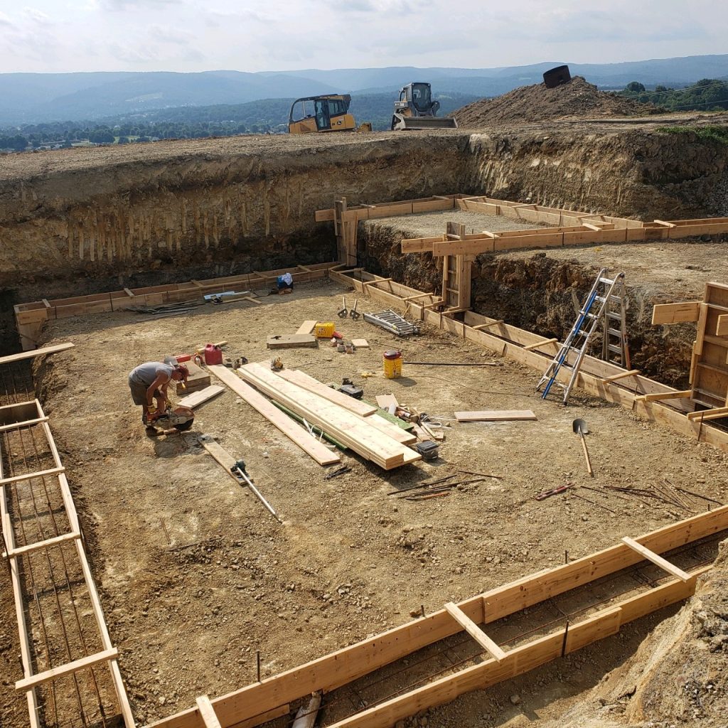 Construction site with workers building wooden forms for concrete foundation on a slope; equipment, tools, and lumber are scattered around the area, with hills visible in the background.