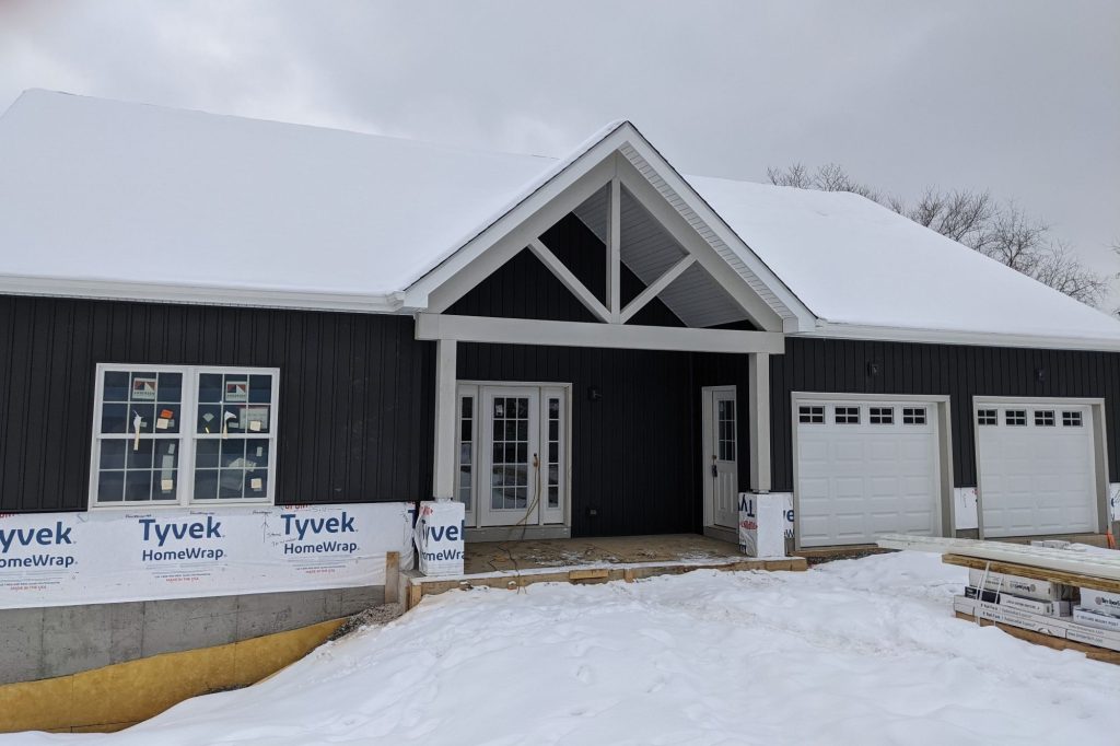 A newly constructed cold climate home design in Pittsburgh with dark siding, white trim, double garage, and a snow-covered roof; construction materials and Tyvek wrap visible.