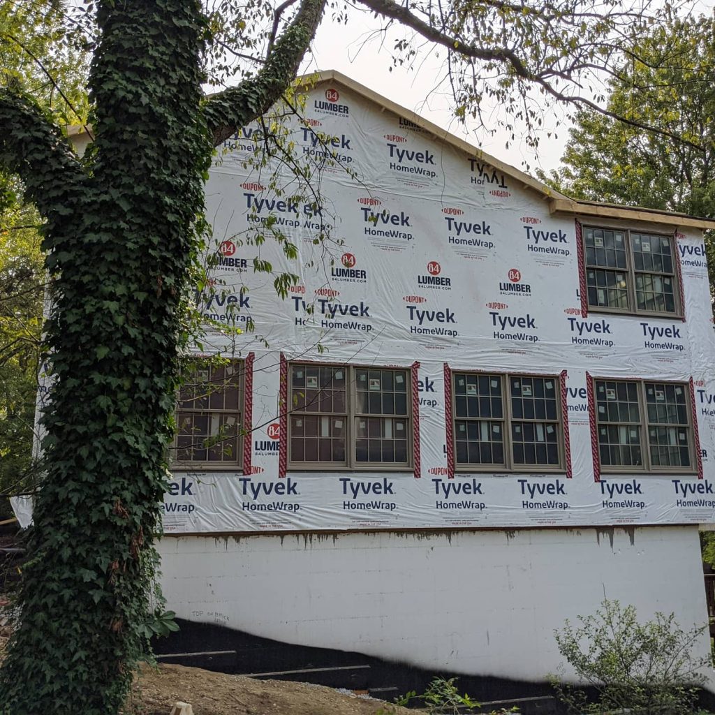 Two-story house under construction with Tyvek HomeWrap covering the exterior walls and multiple windows installed—a great example of cold climate home design; tree with ivy in the foreground.