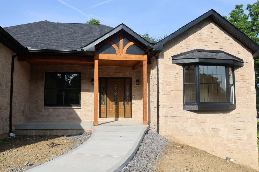 A single-story house with tan brick exterior, black framed windows, and a double wooden front door features a wooden covered entryway—ideal for cold climate home design. A curved concrete path leads to the sheltered entrance.
