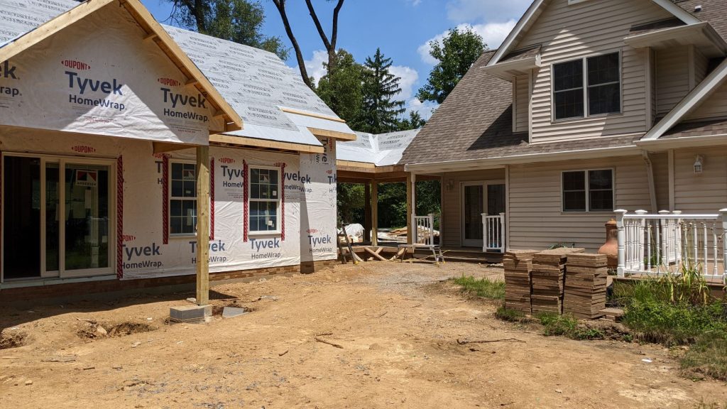 A house under renovation with an unfinished extension covered in Tyvek wrap, stacks of building materials, and exposed soil in the yard, showcasing the possibilities of multi-generational home design.