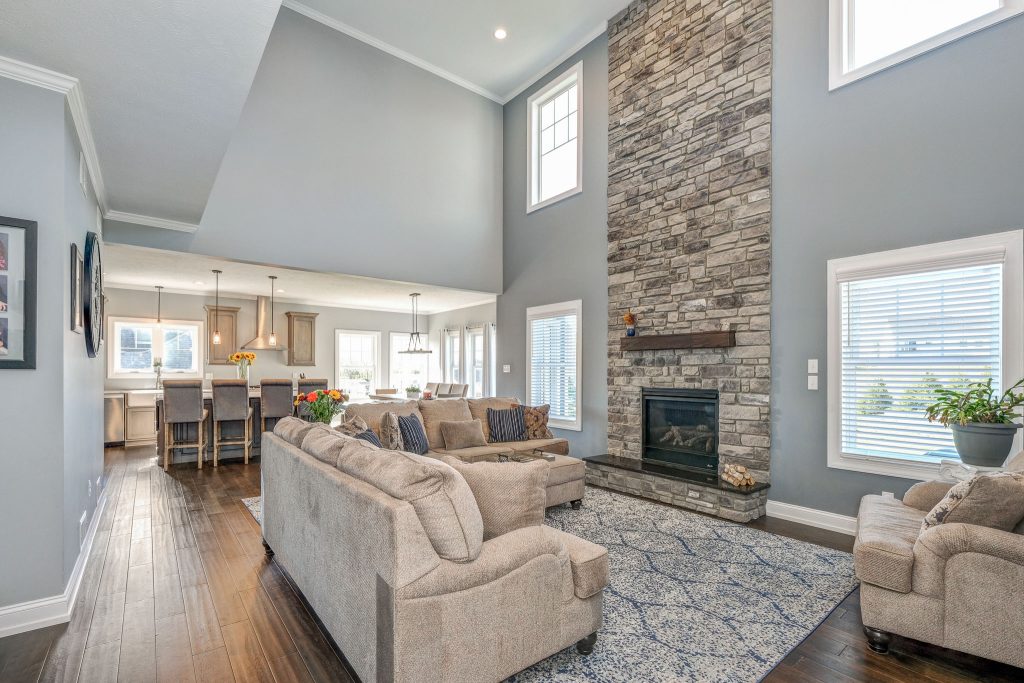 A spacious living room with high ceilings, stone fireplace, gray sofas, and hardwood floors features large windows and an open layout perfect for multi-generational home design, seamlessly connecting the modern kitchen and dining area.