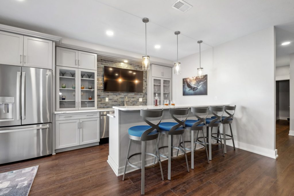 Modern kitchen with stainless steel appliances, an island with four barstools, pendant lighting, and a wall-mounted TV above a tile backsplash—perfect for multi-generational home design and gatherings.