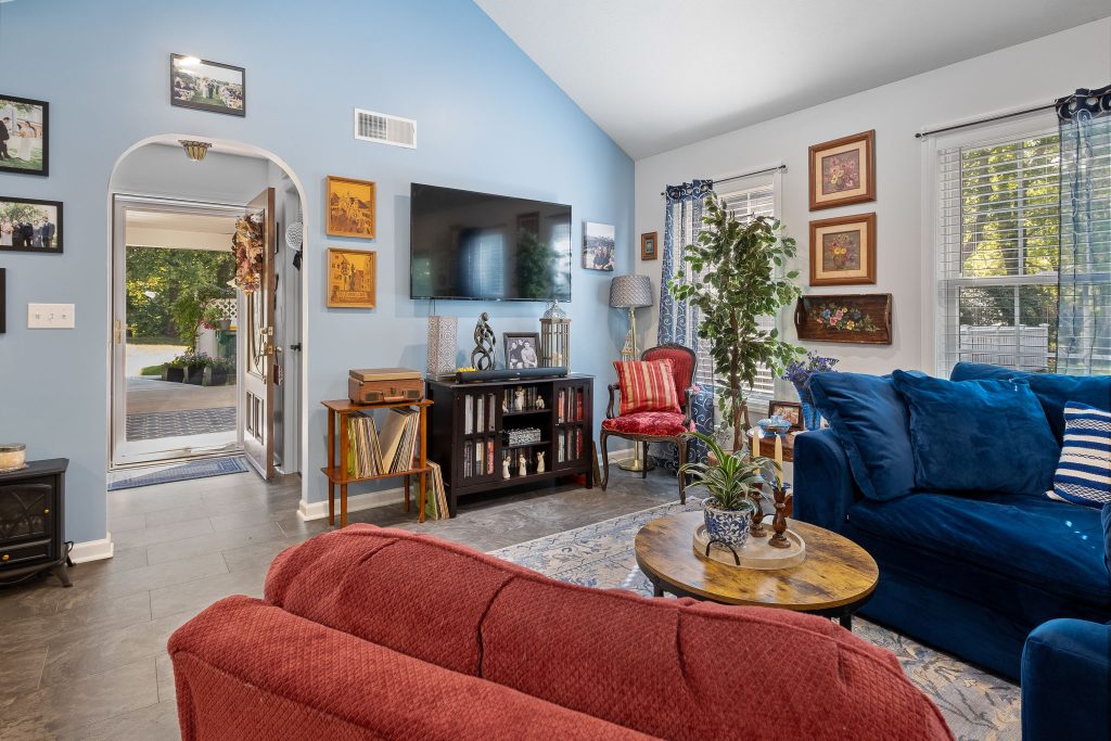 A cozy living room with blue walls, a mounted TV, framed pictures, blue and red couches, houseplants, and large windows fills with daylight—perfect for multi-generational home design that invites everyone to relax together.