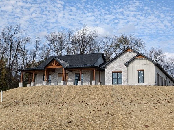 A newly constructed single-story building with stone and wood accents sits on a bare, sloped hill with leafless trees in the background under a partly cloudy sky.