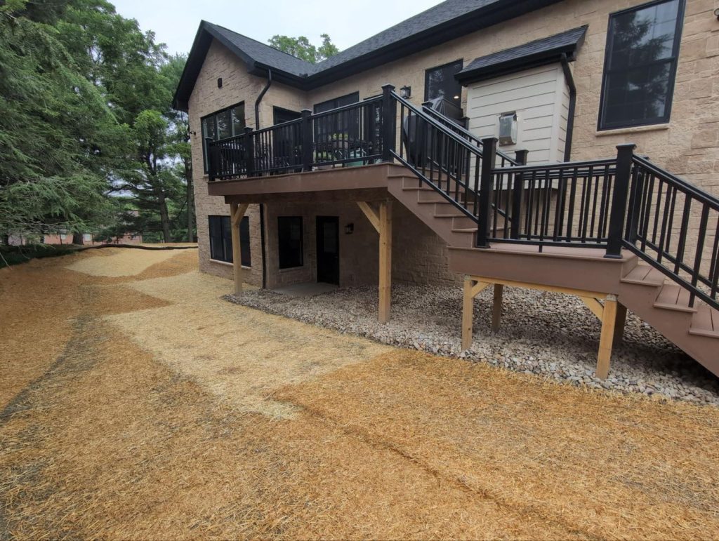 Backyard of a two-story house with a raised deck and stairs, new straw-covered ground, and a mulched area along the side of the house, indicating recent landscaping work.