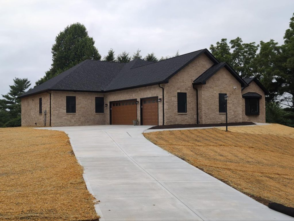 A modern single-story house with tan brick exterior, black roof, three-car garage, and a wide concrete driveway, surrounded by freshly seeded grass and trees in the background.