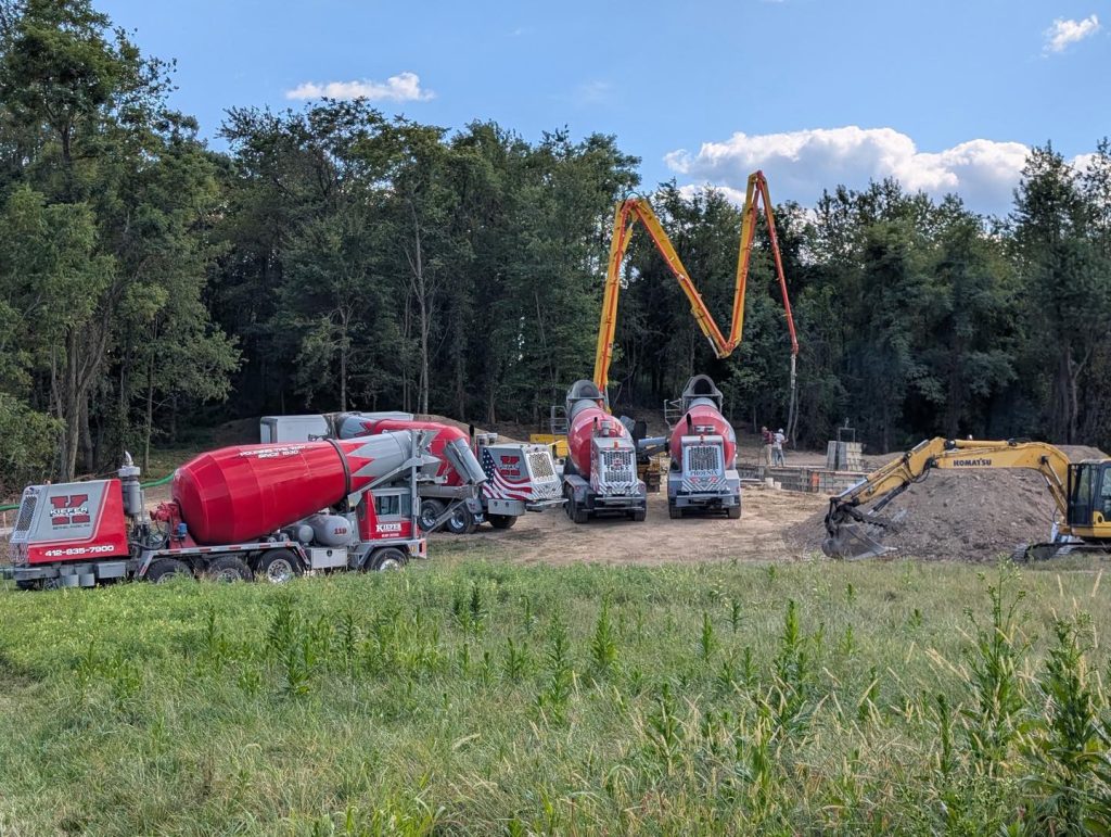 Several cement mixer trucks and a concrete pump operate at a construction site near a wooded area, with an excavator moving dirt in the foreground.