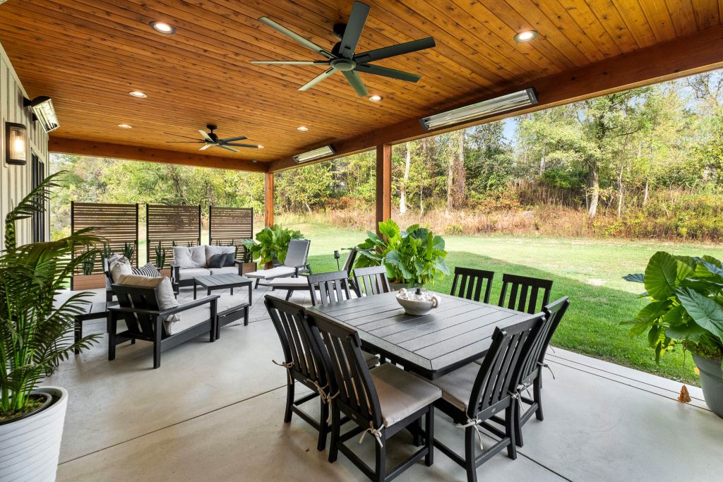 Covered patio with wooden ceiling, ceiling fans, dining table and chairs, outdoor sofas, potted plants, and a view of the grassy yard and trees—showcasing natural materials that are popular in home design trends 2026 for Pittsburgh custom homes.