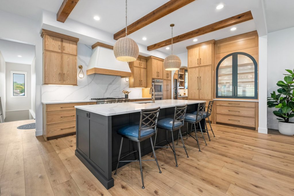 Modern kitchen with light wood cabinets, black island with four barstools, pendant lights, exposed ceiling beams, and a potted plant in the corner highlights natural materials—a hallmark of Pittsburgh custom homes and home design trends 2026.