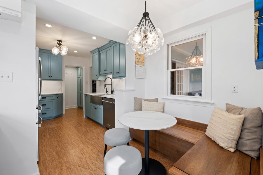 A kitchen with blue cabinets, wood floors, a round table with bench seating, pendant lights, and a window above the bench area highlights natural materials and reflects top home design trends for 2026 in Pittsburgh custom homes.