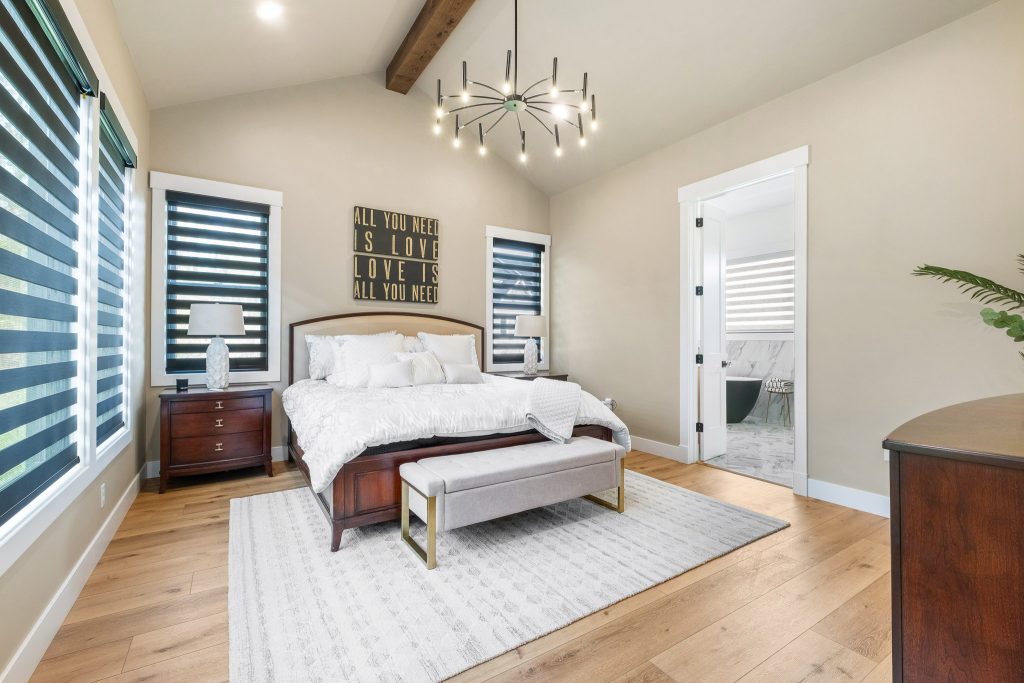 A modern bedroom in the style of Pittsburgh custom homes features a wooden bed, white bedding, nightstands, a bench, and large windows; a bathroom is visible through an open door.