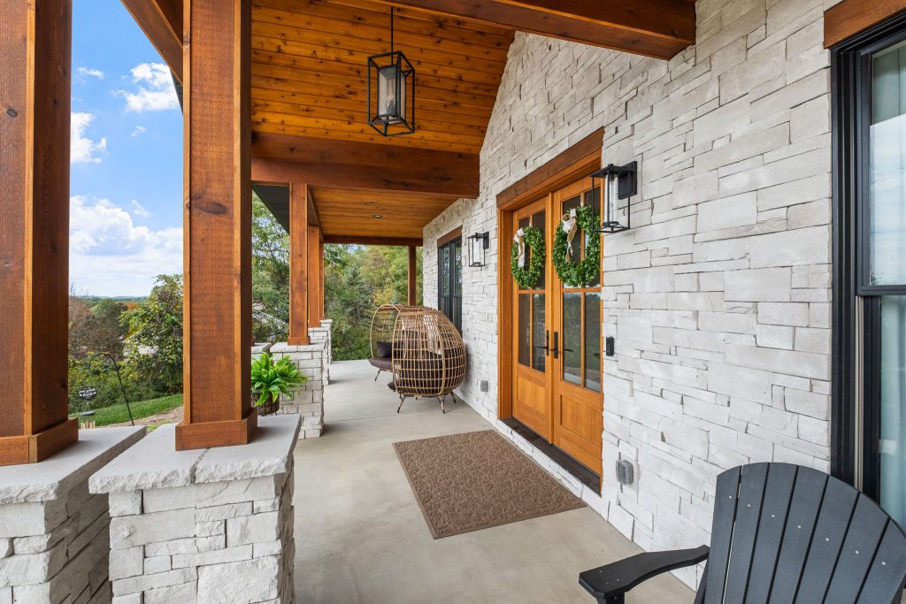 A covered porch with white stone walls, wooden double doors, black outdoor chairs, a brown hanging wicker chair, and green plants showcases natural materials perfect for Pittsburgh custom homes and upcoming home design trends 2026.