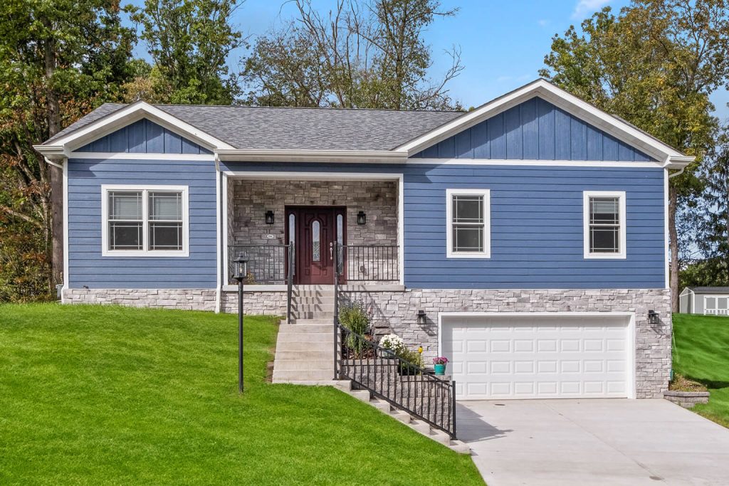 Single-story house with blue siding, stone accents, a front porch, and an attached two-car garage. Designed by a custom home builder Pittsburgh residents trust. Grassy lawn and trees in the background; steps and a ramp lead to the entrance.