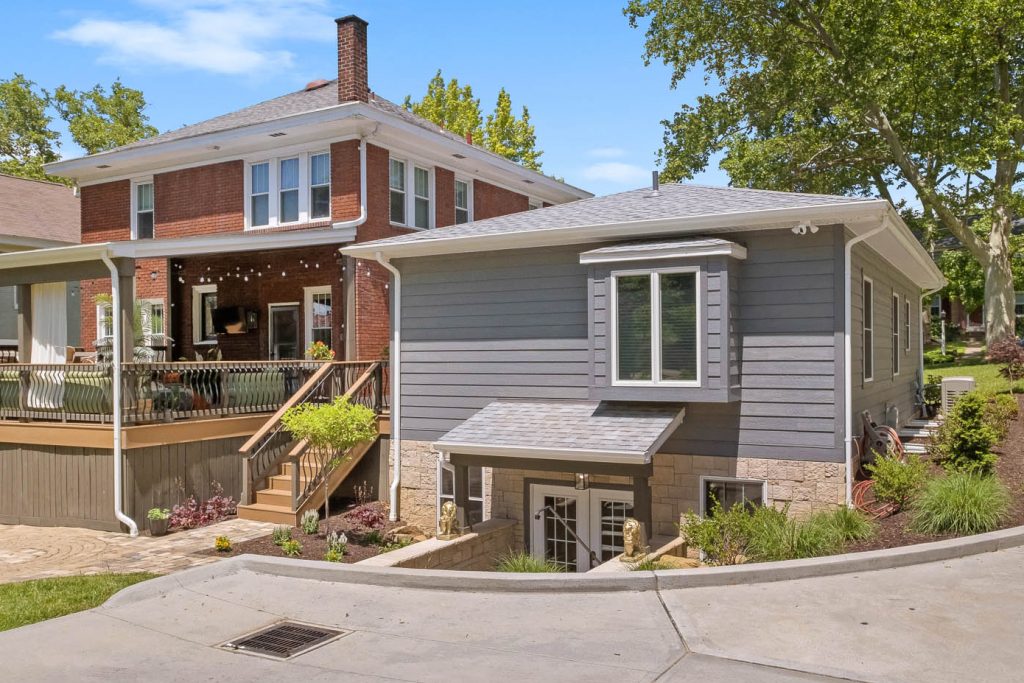 A gray single-story house with a basement entrance sits next to a red brick two-story home, both surrounded by trees and landscaping—showcasing potential for stylish home additions Pittsburgh residents may love.